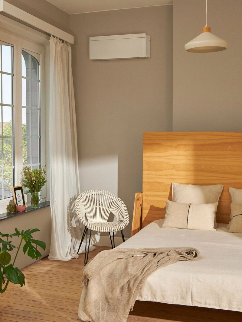 A wall-mounted air-to-air heat pump indoor unit installed high on the wall of a bright bedroom with a bed, chair and large window