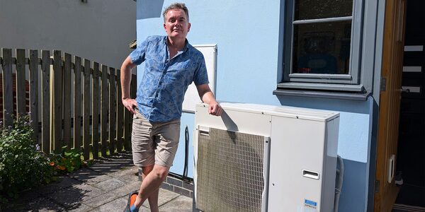 A man standing beside a white outdoor heat pump unit on the exterior wall of a house