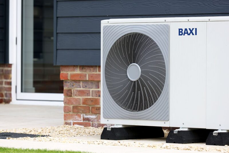 A white Baxi air source heat pump monobloc unit installed outside a house on black mounting feet beside a brick wall and dark cladding