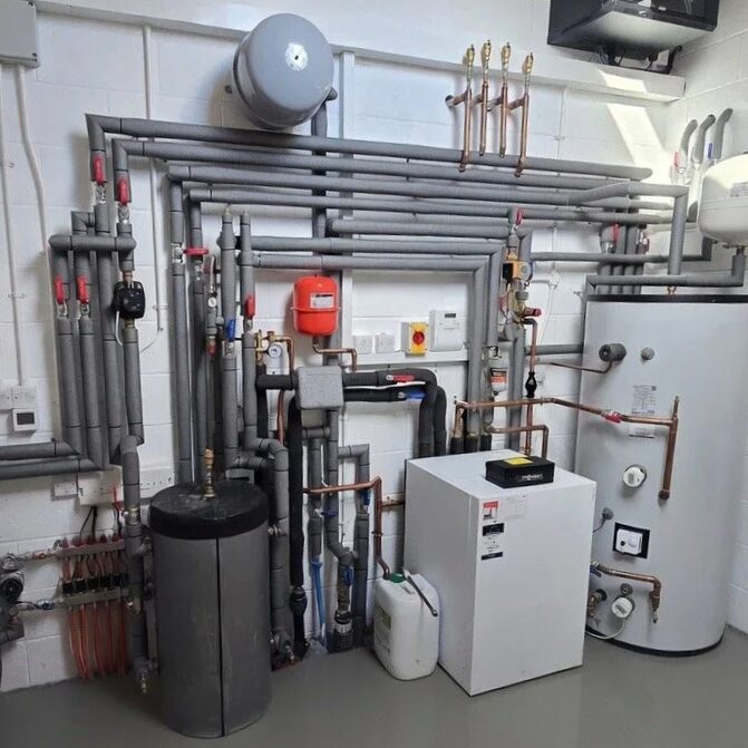 A heat pump plant room installation with a white heat pump unit, hot water cylinder, buffer vessel, expansion vessels and extensive grey and copper pipework on a white brick wall