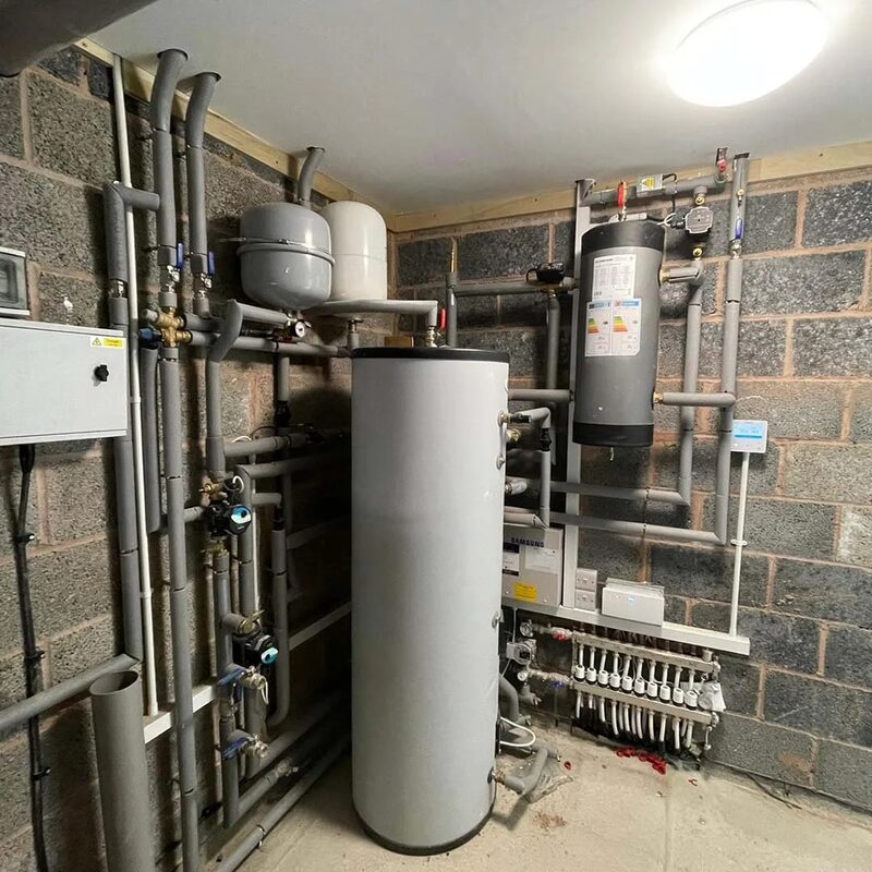 A heat pump heating system installation with a large white hot water cylinder, buffer vessel, expansion vessels, manifold and grey pipework in a plant room with block walls