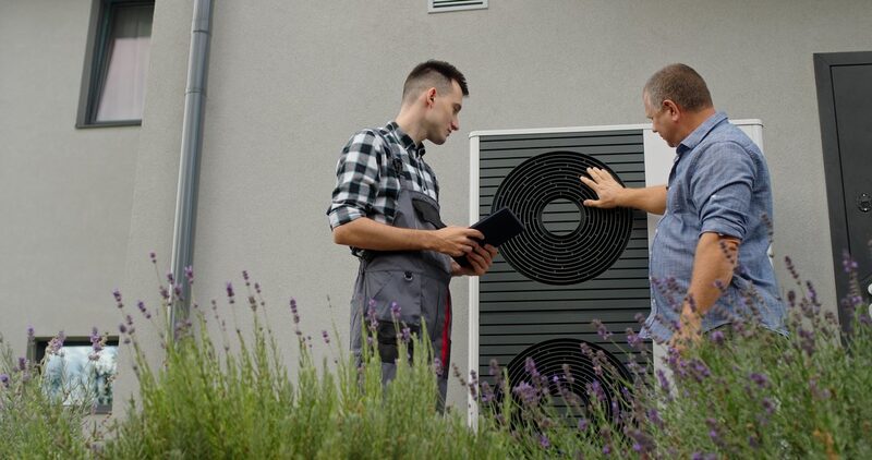 An installer in overalls holding a tablet discussing an air source heat pump unit outside a house with a homeowner