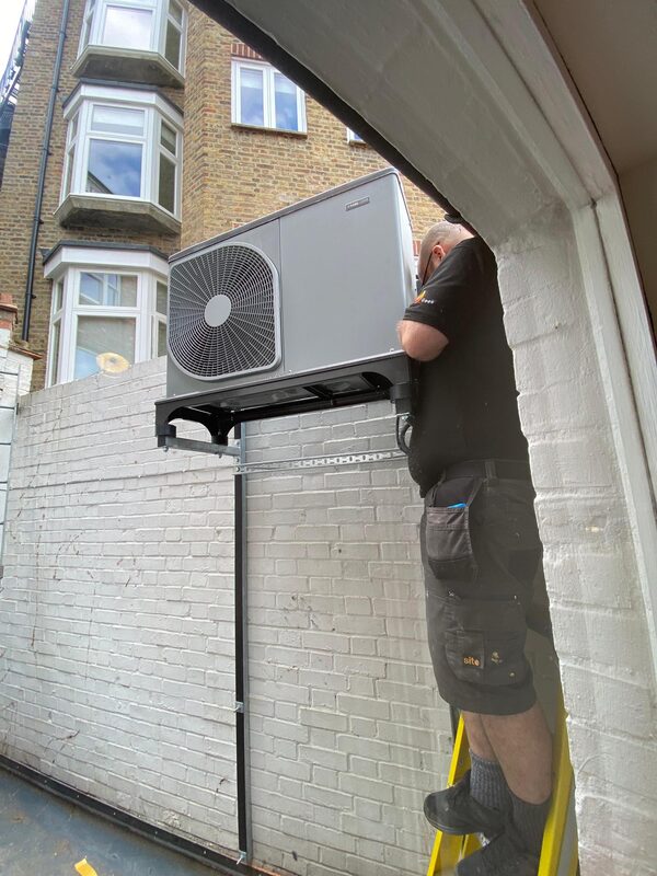 An engineer on a ladder installing a wall-mounted NIBE air source heat pump unit on a white brick wall outside a UK home