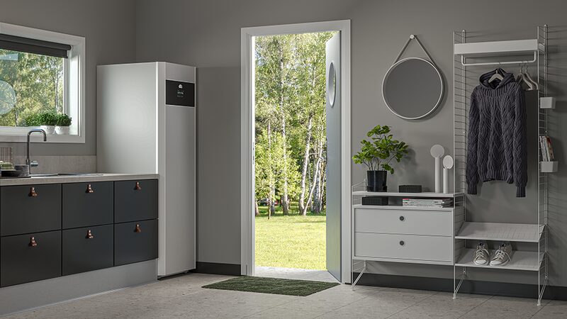 A white indoor heat pump / hot water cylinder unit installed in a grey utility room beside kitchen cabinets and an open door to a garden