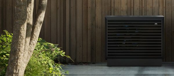 A dark grey outdoor air-to-water heat pump unit installed against a wooden fence beside a tree and shrubs