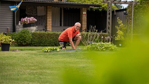 A man in an orange shirt crouching in a garden beside a ground source heat pump collector loop in the lawn, with a house behind