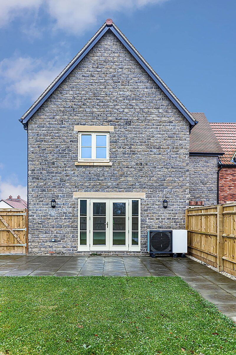 A heat pump outdoor unit installed on a paved patio beside the rear of a stone house with a lawn and wooden fencing