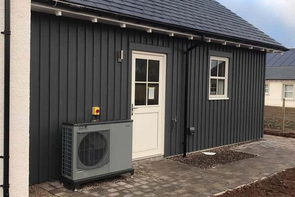 An air source heat pump unit installed outside a dark grey timber-clad house with a white door