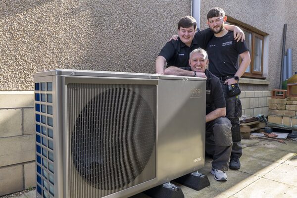 Three installers posing outdoors beside an Ideal HP290 air source heat pump installed against a house wall
