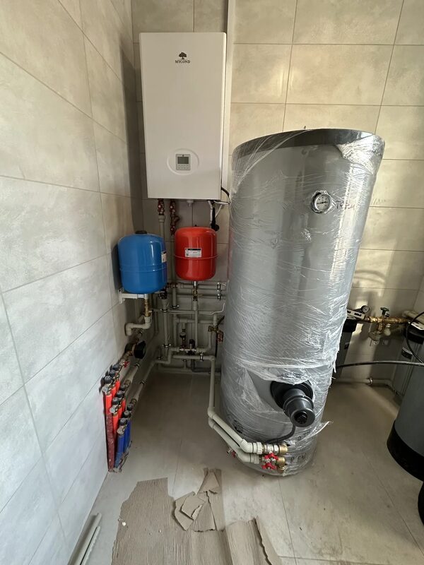 A wall-mounted Mycond heat pump indoor unit with expansion vessels, pipework and a large wrapped hot water cylinder in a tiled utility room