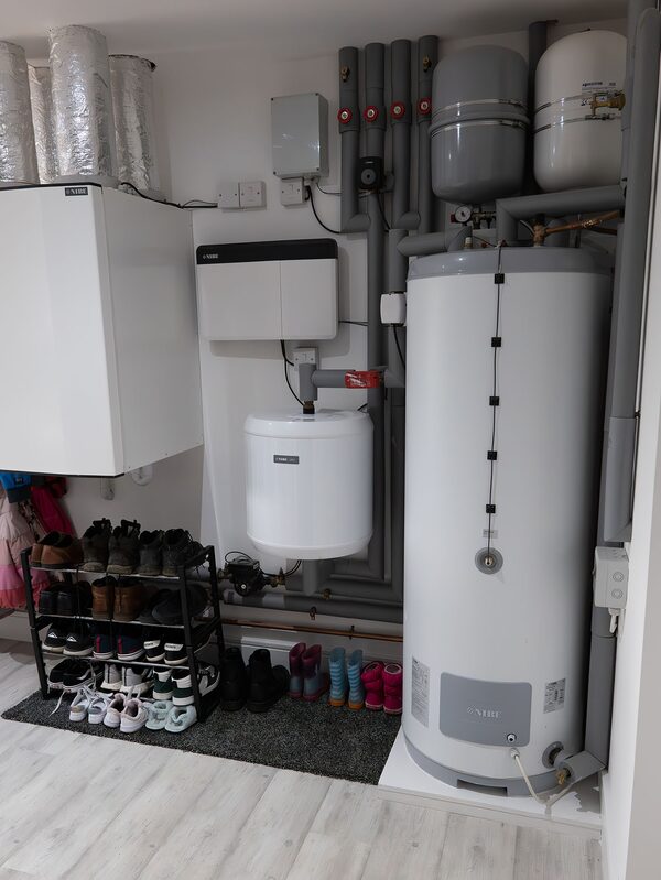 A NIBE heat pump indoor unit with hot water cylinder and buffer vessels installed in a home utility space, with a shoe rack of family footwear nearby