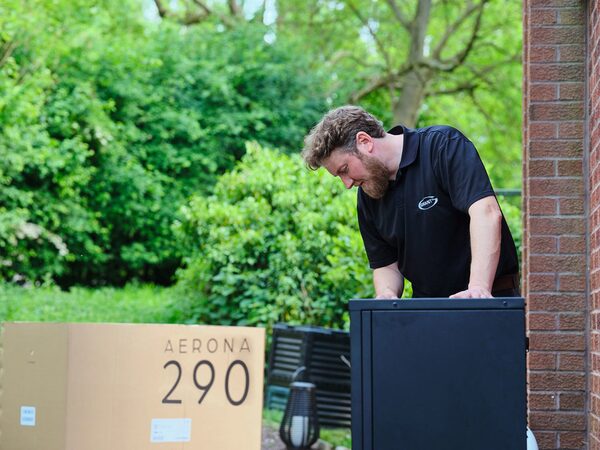 An installer in a branded polo shirt unpacking a Grant Aerona 290 heat pump unit outside a brick house