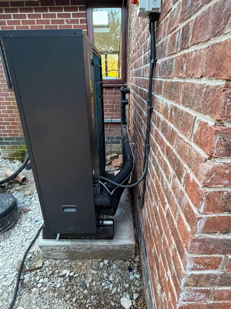 A Grant air source heat pump unit installed on a concrete slab against a brick house wall with insulated pipework running to the property.