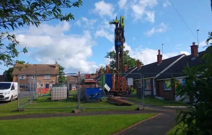 A ground source heat pump borehole drilling rig operating behind temporary fencing on a grassy area between brick bungalows on a housing estate