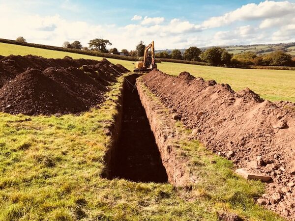 A long excavated trench in a grassy field with a yellow digger, dug for ground source heat pump pipework