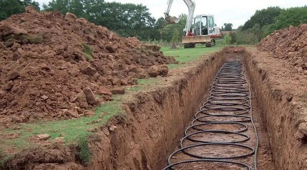 A long excavated trench with coiled black ground-loop pipework laid inside, with an excavator and soil heaps in the background