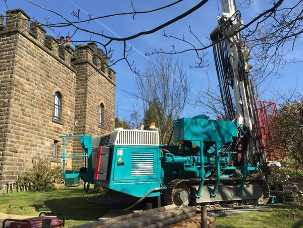 A tracked drilling rig boring a ground-source borehole in the garden of a stone castle-style house