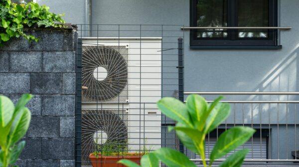 An air source heat pump unit with two fans installed outside a grey house behind a metal guard railing