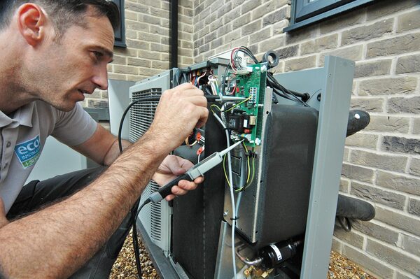 A technician testing the wiring and control board of an outdoor heat pump unit outside a brick house