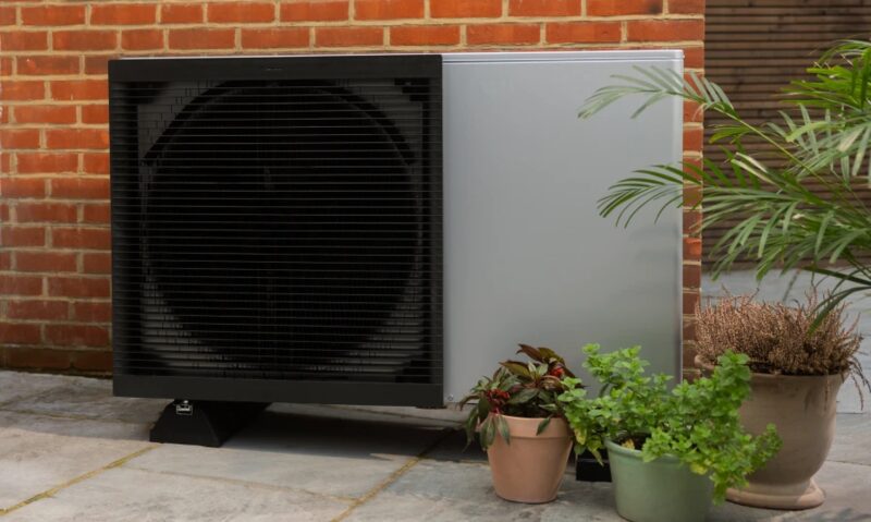 An air source heat pump unit installed outside a brick house, with potted plants beside it on a paved patio