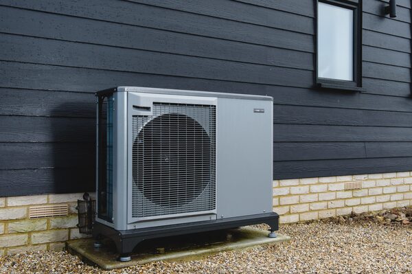 A grey air source heat pump unit installed outside a dark-clad house on a gravel bed