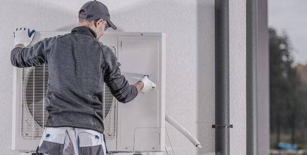 A technician in work clothes installing an outdoor air source heat pump unit on an exterior wall