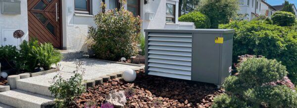 An outdoor air-source heat pump unit installed beside the front entrance of a house with a garden