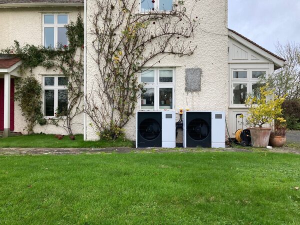 Two air source heat pump units installed side by side on the exterior wall of a cream-rendered house with a lawn in front