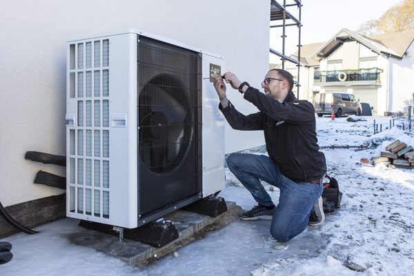An engineer kneeling in the snow working on an air source heat pump unit outside a house
