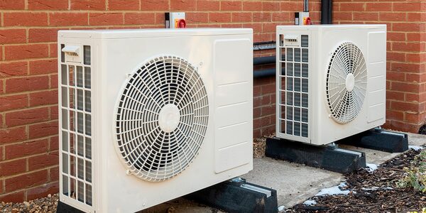 Two white air source heat pump units installed outside against a red brick house wall