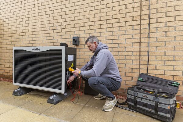 A technician using a multimeter to service an outdoor HISA air source heat pump beside a brick wall, with an open tool bag nearby