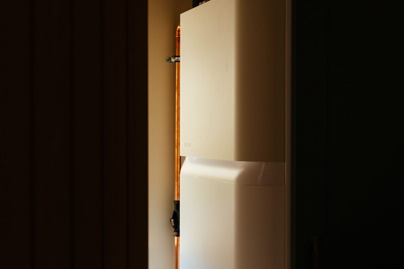 A white indoor heat pump or hot water cylinder unit installed in a dark cupboard with copper pipework beside it