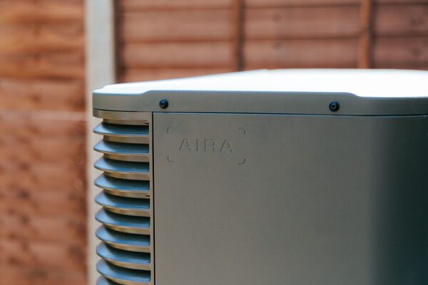 A close-up of a grey Aira air source heat pump unit installed outdoors against a wooden fence