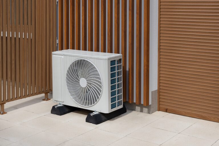 An air source heat pump outdoor unit installed on anti-vibration mounts on a paved patio beside timber slat fencing