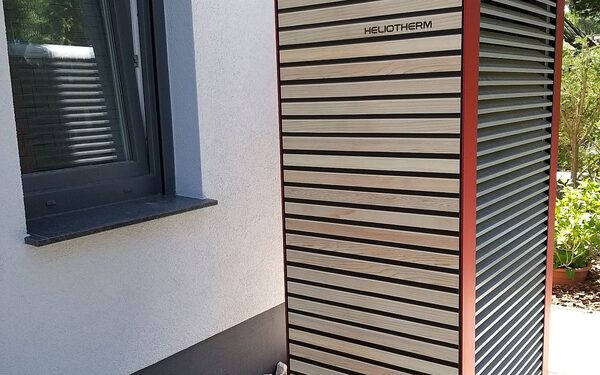 A Heliotherm air-source heat pump unit with timber slat cladding installed outside a house wall next to a window