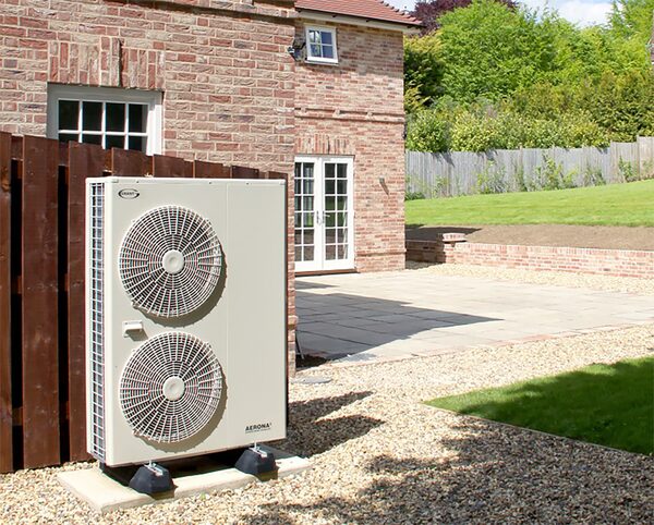 A Grant Aerona air source heat pump unit installed outside a brick house behind a wooden fence on a gravel base