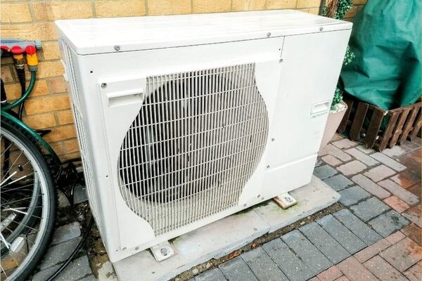 A white air source heat pump unit installed outside a brick house on a paved patio