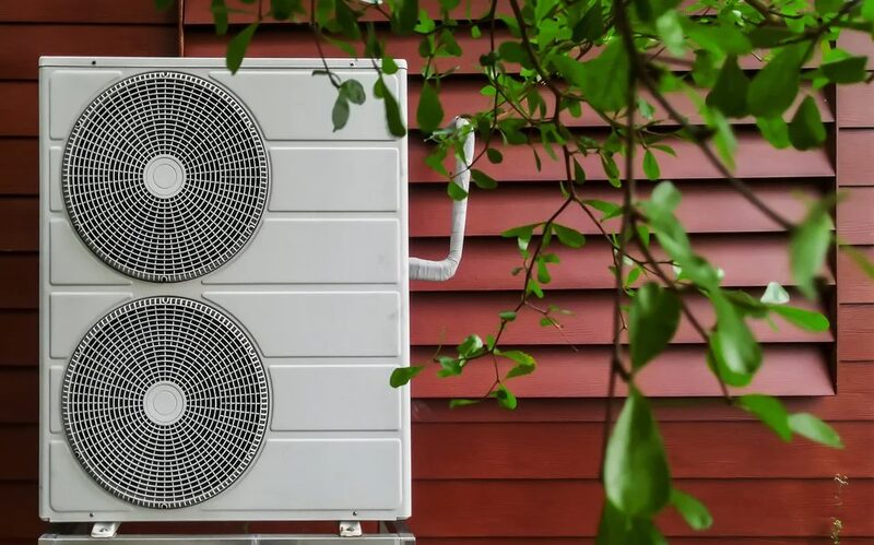An air source heat pump unit with two fans mounted on the outside wall of a red wooden-clad house, partly framed by green leaves