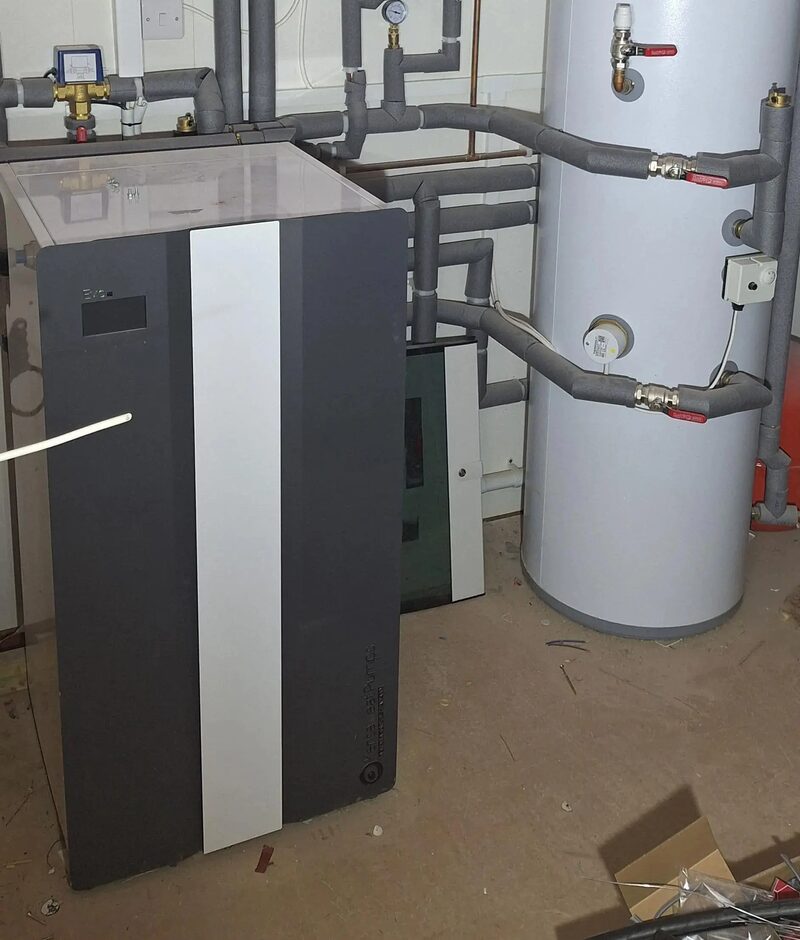 A Kensa ground source heat pump unit installed indoors next to a hot water cylinder with insulated pipework