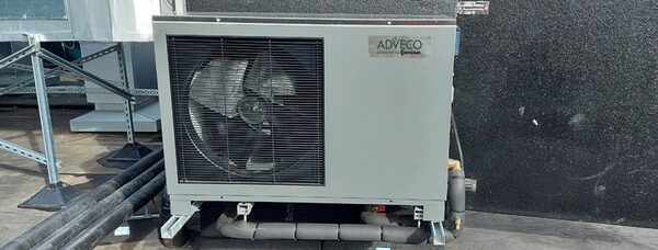 An Adveco-branded air source heat pump unit with a large fan installed outdoors on a flat roof next to pipework