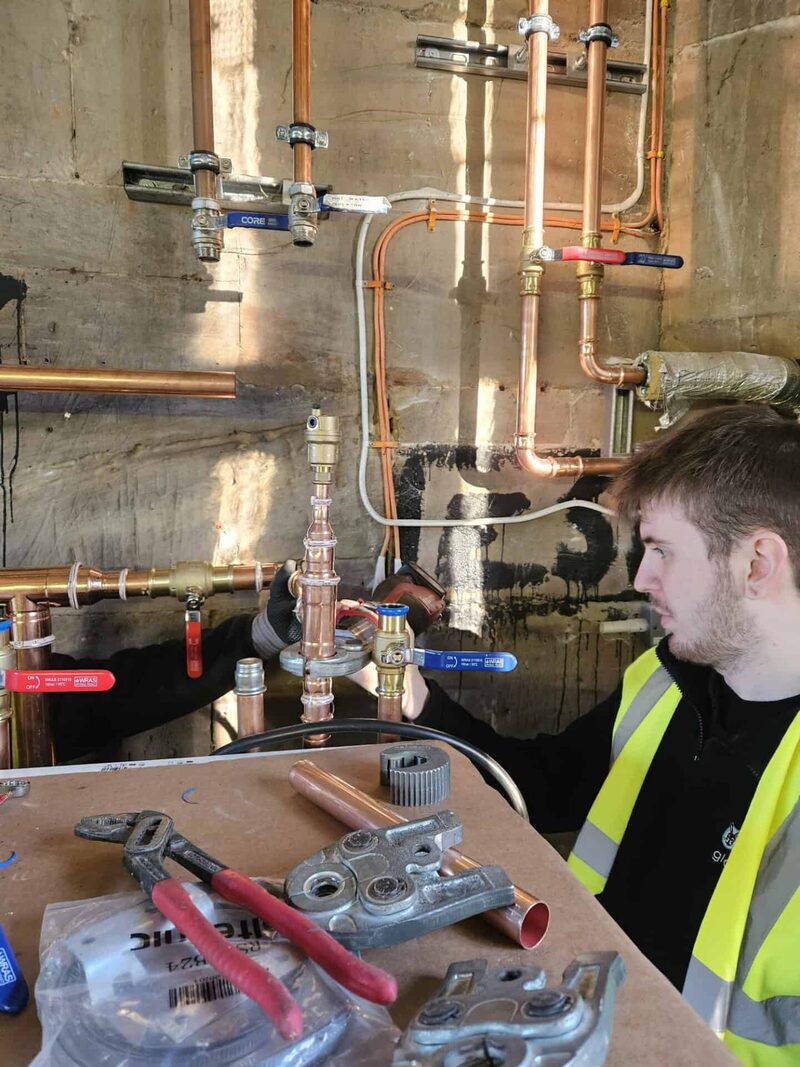 A heating engineer in a high-visibility vest working on copper pipework and valves for a ground source heat pump installation