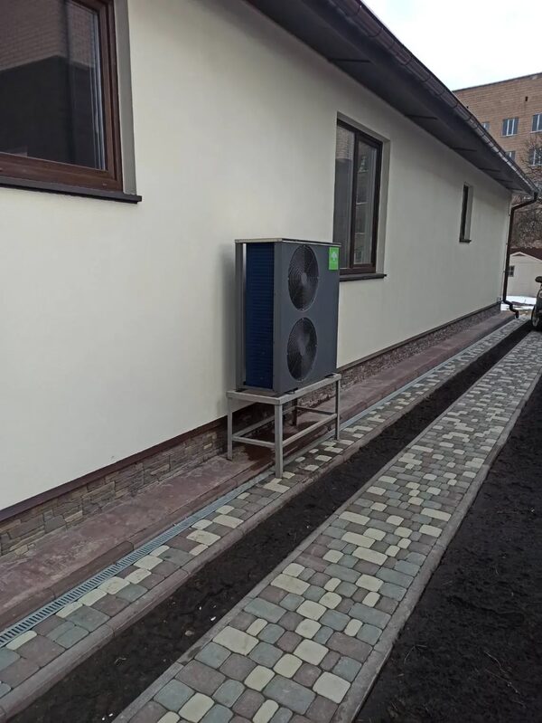 An air source heat pump unit installed on a metal stand outside a cream-rendered private house beside a block-paved path