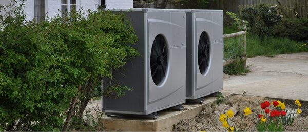 Two grey air source heat pump units installed outside a house beside a garden path and flower bed