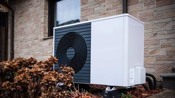 An air source heat pump unit installed outside a brick house, partially hidden by dried plants