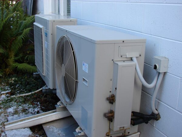 Two heat pump outdoor units installed against the white brick wall of a house with pipework and cabling