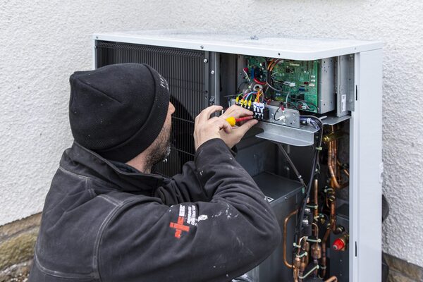 An engineer in a black beanie and jacket working on the open electrics of an outdoor air source heat pump unit against a white wall