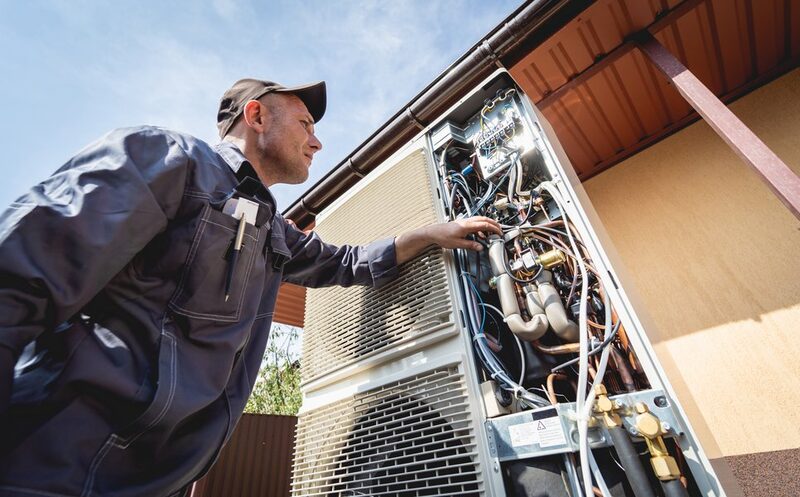 A technician in work overalls inspecting the open internal components of an outdoor heat pump unit beside a house