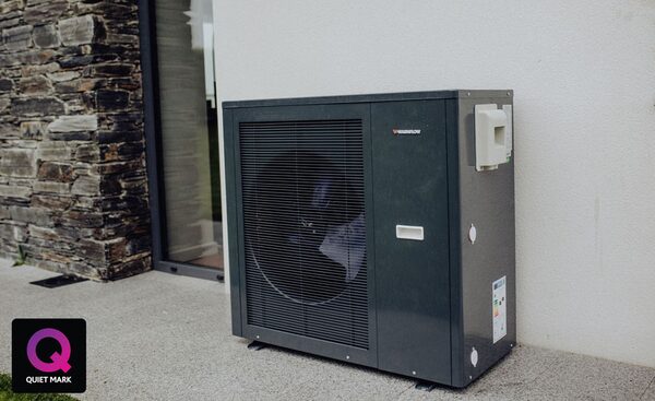A dark grey air source heat pump unit installed outside a house next to a stone wall and glass door