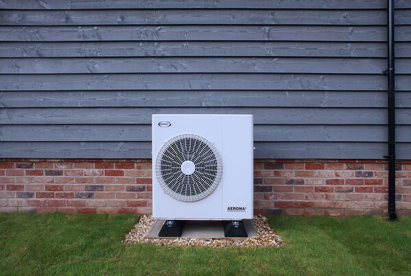 A Grant Aerona air source heat pump unit installed outside a house against a grey weatherboard and brick wall