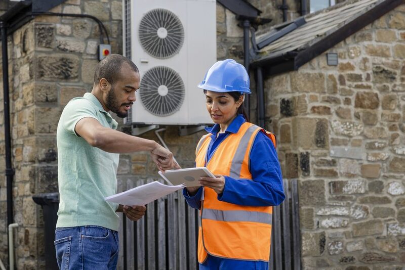 A heat pump installer in a hard hat and hi-vis vest discusses paperwork with a householder outside a stone house with an air source heat pump unit on the wall